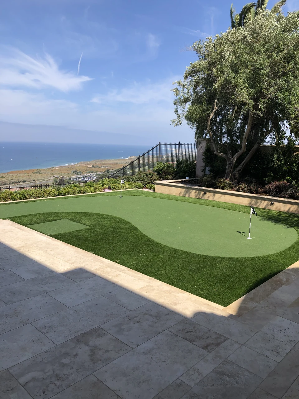 Putting green with flagsticks and a darker turf fringe beside a stone terrace overlooking the coast