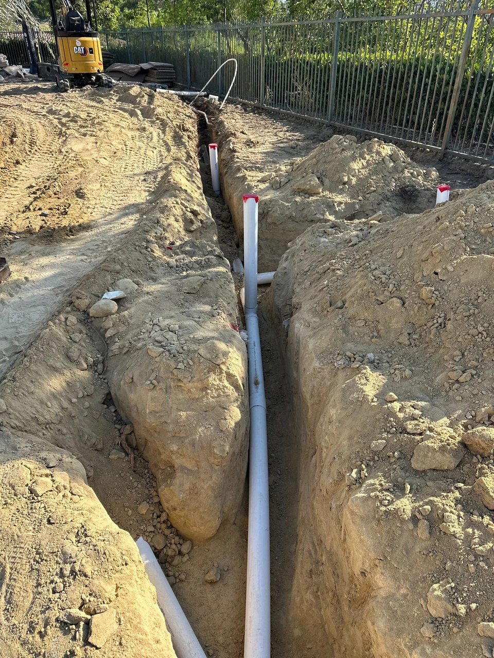 Gravity-drain piping with upright connections in a trench beside a fenced yard