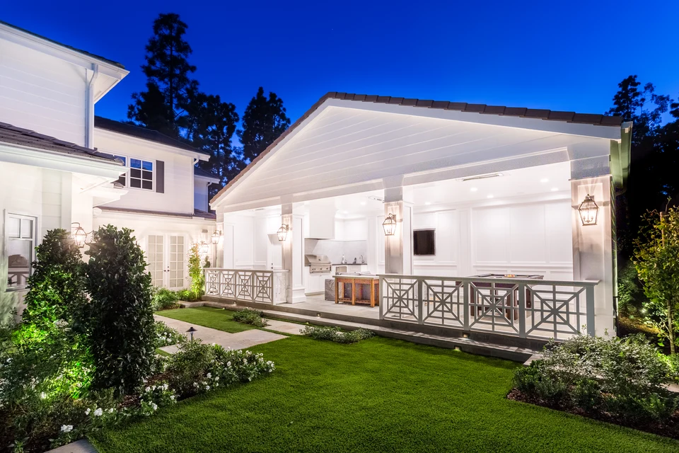 Illuminated white pool house and adjoining home at dusk with lanterns and garden lighting