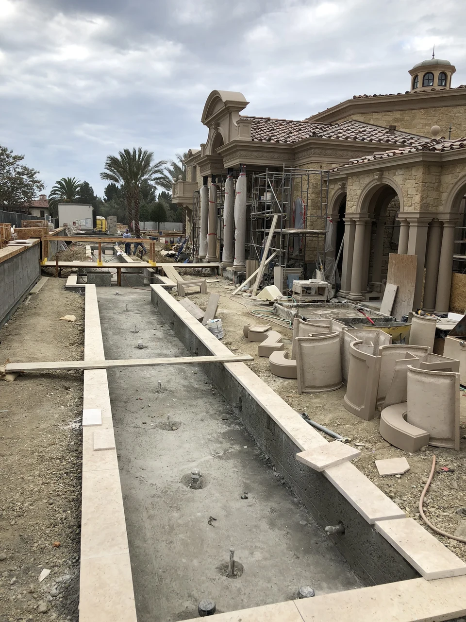 Long water-feature basins with pale stone edges under construction beside a large residence