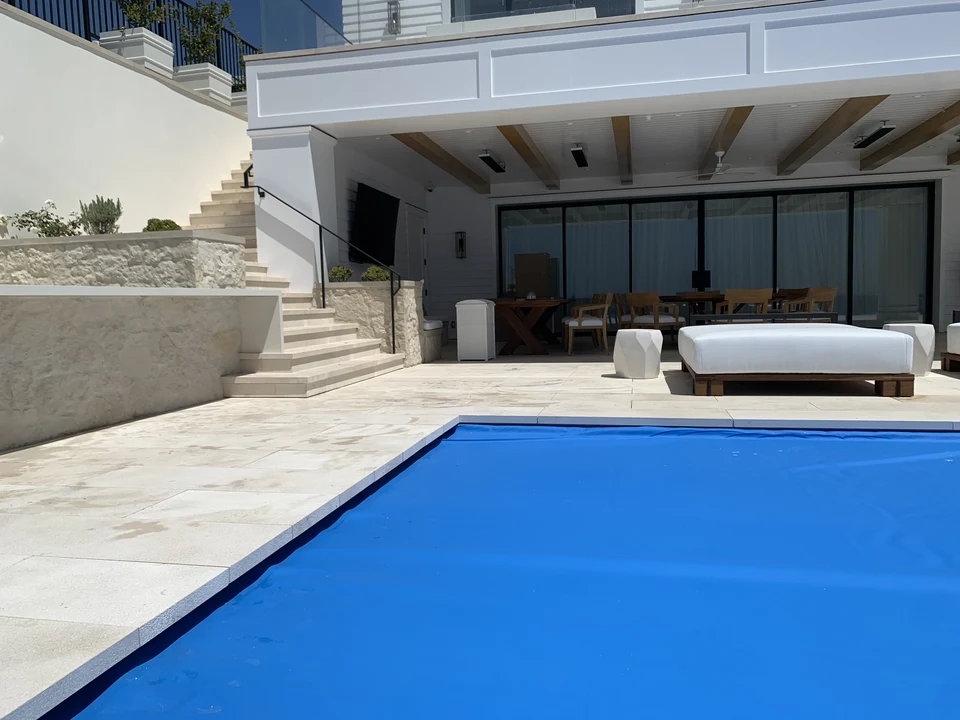 Pale terrace paving beside a covered swimming pool, shaded seating and a stone stairway
