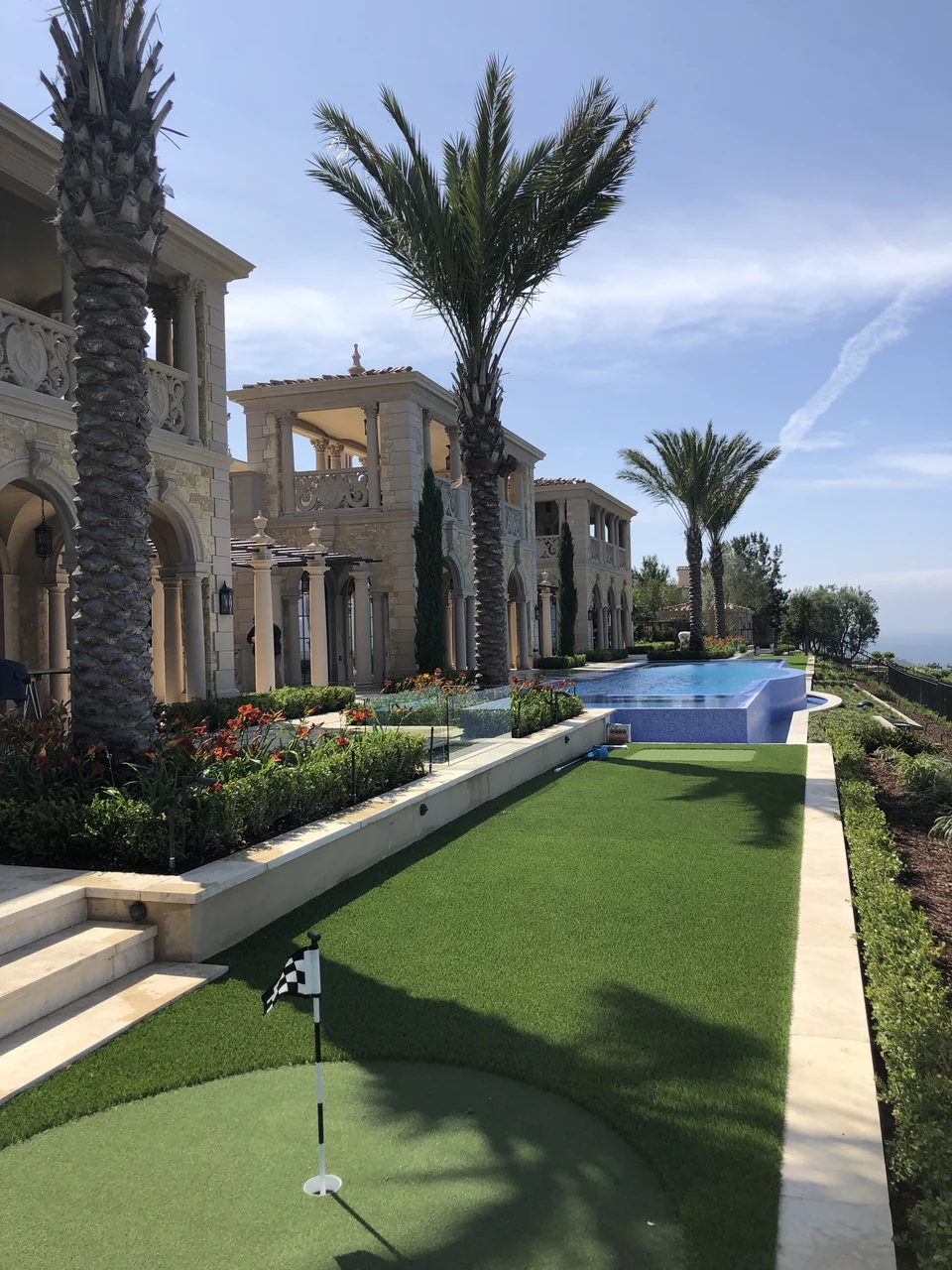 Putting-green flag in the foreground with a long artificial lawn, palm trees and pool beyond